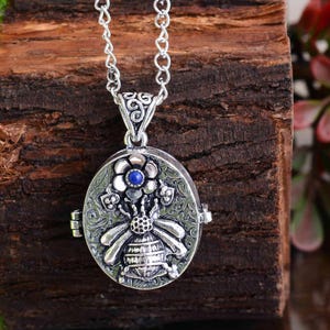 Puede incluir: Collar con colgante ovalado plateado con diseño de abeja y flor. El relicario presenta una abeja detallada en el centro, rodeada de elementos florales y decorativos. Una pequeña piedra azul está engastada en el centro de la flor. El collar tiene una cadena delicada.