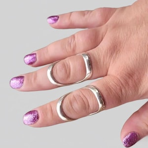May include: Two silver-colored finger rings with a rectangular design. The rings are worn on the index and middle fingers. The nails are painted with a sparkly purple nail polish.