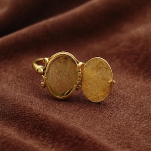 May include: A gold locket ring with an oval-shaped compartment that opens to reveal a small space. The ring is set against a brown background. The locket is open, showing the interior.