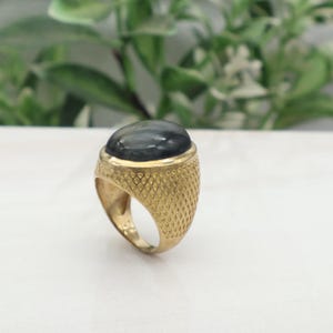 May include: A gold-colored ring with a large, oval, dark gray gemstone. The ring has a textured, diamond-patterned band. The gemstone is set in a smooth bezel. The ring is displayed on a white surface with a blurred green background.
