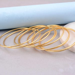Könnte beinhalten: Eine Sammlung goldfarbener Armreifen, leicht überlappend angeordnet. Die Armreifen sind dünn und kreisförmig und reflektieren das Licht. Der Hintergrund zeigt eine hellblaue und graue Oberfläche mit einem weißen Stein. Diese Armreifen sind eine Art Schmuck.