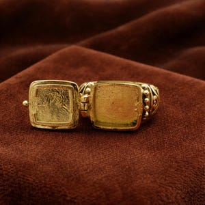 May include: A gold-colored locket ring with a hinged square compartment. The ring is open, revealing the interior. The band features decorative beading and scrollwork. The ring is set against a brown background.