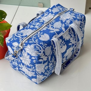 May include: A blue and white floral patterned duffel bag. The bag features a zipper closure, two handles, and a detachable shoulder strap. The floral design is a repeating pattern of white flowers and leaves on a blue background.