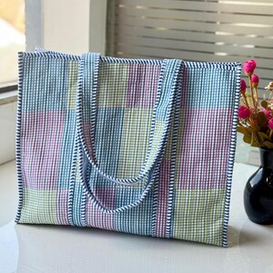 May include: A colourful tote bag with a plaid pattern in shades of blue, green, and pink. The bag has matching handles and trim. It is made of a woven fabric and sits on a white surface. The bag is rectangular.