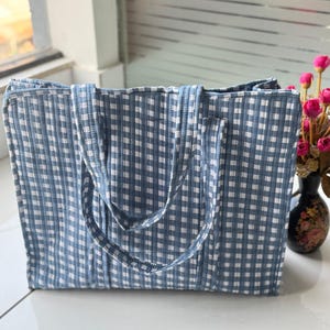Może przedstawiać: Niebiesko-biała torba typu tote w kratę gingham z pasującymi uchwytami. Torba jest prostokątna i wydaje się być wykonana z pikowanej tkaniny. Torba stoi na białej powierzchni.