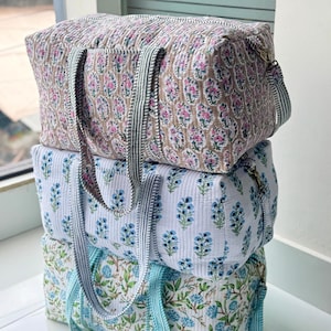 Puede incluir: Tres bolsas de viaje acolchadas apiladas. La bolsa superior es de color canela con estampados florales rosas y blancos. La bolsa del medio es blanca con estampados florales azules. La bolsa inferior es blanca con estampados florales azules y verdes. Cada bolsa tiene asas a rayas.