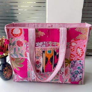 Könnte beinhalten: Gesteppte Tragetasche mit Patchwork-Design. Die Tasche hat eine Mischung aus Rosa und floralen Mustern, mit weißen Verzierungen und Griffen. Sie hat eine rechteckige Form und eine kleine Tasche auf der Vorderseite.