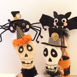 May include: Three Halloween decorations: a black spider with a clock face on its hat, a black bat with pink cheeks and a toothy grin, and a white skull wearing a black top hat with a striped band and a clock face on the hat.