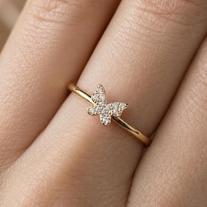 Op de afbeelding: Een gouden ring met een vlindermotief. De vlinder is versierd met kleine, sprankelende steentjes. De ring wordt getoond op een vinger, waarbij de fijne details van het sieraad worden benadrukt.