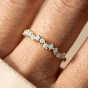May include: A gold ring featuring a row of small, round opals. The opals have a milky white base with flashes of blue and green. The ring is worn on a finger, showcasing the delicate design and craftsmanship.