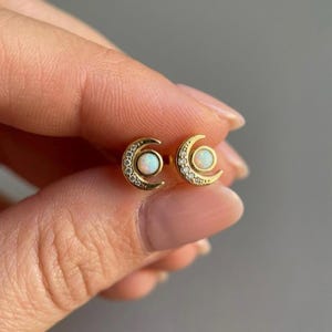 May include: A pair of gold-colored crescent moon stud earrings. Each earring features a central, round, white opal stone, surrounded by small, clear crystals. The earrings are designed with a celestial theme.