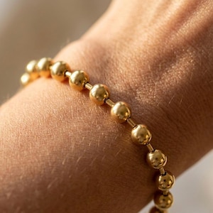 May include: A gold-colored beaded bracelet. The bracelet features a series of round, polished beads strung together. The beads are evenly spaced and have a smooth, reflective surface. The bracelet is worn on a wrist, with the skin tone visible.