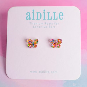 Peut inclure: Boucles d'oreilles papillon avec des accents irisés colorés. Elles sont présentées sur une carte blanche avec le nom de la marque "aidille" et le texte "Titanium Posts for Sensitive Ears". Fond rose et bleu pastel.