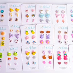 May include: A collection of stud earrings on white cards. The earrings feature various designs, including animals, food, and floral patterns. Colors include blue, yellow, pink, and green. The cards have the brand name "ainlue" printed on them.