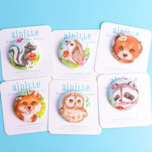 May include: A collection of six round magnets featuring watercolor illustrations of woodland animals, including a skunk, hedgehog, bear, fox, owl, and raccoon. Each magnet is mounted on a white card with the brand name "aidille".