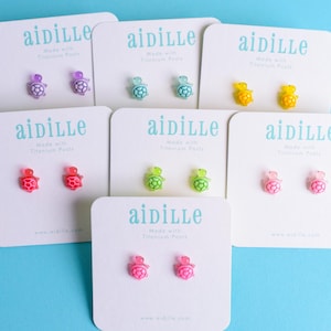 May include: Seven pairs of turtle-shaped stud earrings in purple, teal, yellow, red, green, light pink, and hot pink. Each pair is mounted on a white card labeled with the brand name aidille and the text Made with Titanium Posts.