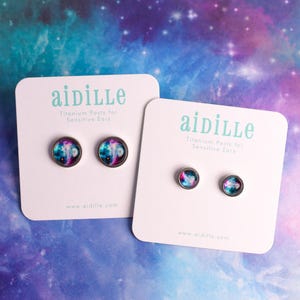 May include: Two pairs of round stud earrings with a galaxy design are displayed on white cards. The cards have the brand name "aidille" and the text "Titanium Posts for Sensitive Ears". The background is a colorful galaxy pattern.