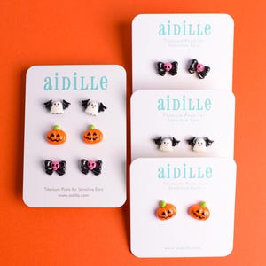 May include: Halloween-themed stud earrings on white cards against an orange background. The earrings feature designs of ghosts, pumpkins, and bows with skulls. The cards display the brand name "aidille".