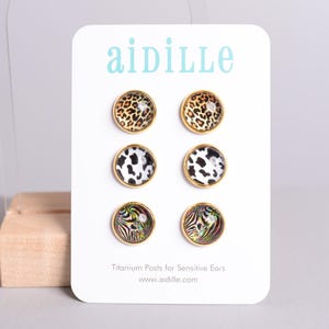 May include: A set of six stud earrings with gold-colored rims, displayed on a white card. The earrings feature animal print designs, including leopard, cow, and zebra patterns. The card has the brand name "aidille" and the text "Titanium Posts for Sensitive Ears".