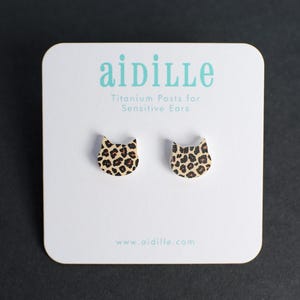 May include: A pair of cat-shaped stud earrings with a leopard print design. The earrings are displayed on a white card with the brand name "aidille" and the text "Titanium Posts for Sensitive Ears". The website address www.aidille.com is also visible.