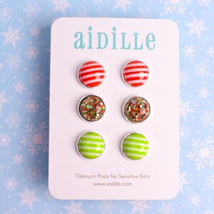 May include: A set of six stud earrings on a white card with the brand name "aidille". The earrings have silver-colored metal settings. The top row features red and white striped earrings. The middle row has glittery earrings. The bottom row has green and white striped earrings.