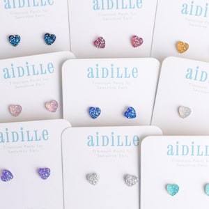 May include: Nine pairs of heart-shaped stud earrings with glitter on a white background. The earrings are in various colors, including blue, pink, purple, silver, and yellow.