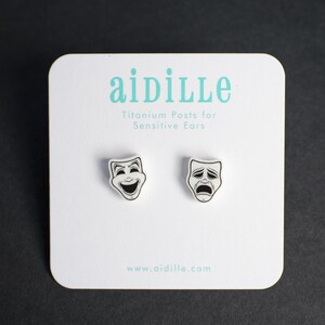 May include: A pair of silver-colored drama mask earrings, one smiling and one frowning, on a white card. The card has the text "aidille" in teal, with "Titanium Posts for Sensitive Ears" below. The website address www.aidille.com is at the bottom.