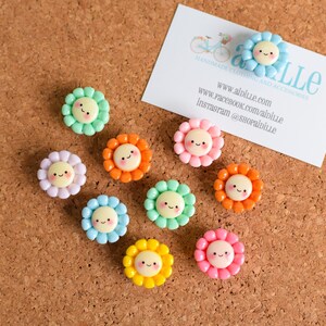 May include: A collection of ten small, circular flower-shaped buttons in various pastel and bright colors including mint, lavender, orange, blue, yellow, and pink. Each button features a central cream-colored face with a simple smiling expression.
