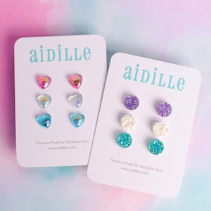 May include: Two white cards with the brand name "aidille" printed in light blue. Each card displays three pairs of stud earrings in various colors and shapes, including hearts and round studs. The text "Titanium Posts for Sensitive Ears" and website address are also visible.