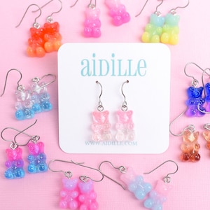 May include: Assortment of colorful gummy bear dangle earrings. Each pair features two gummy bears stacked on top of each other. The colors include pink, blue, green, orange, purple, and yellow. The earrings are displayed on a pink background with a white card that says "aidille" and "www.aidille.com".