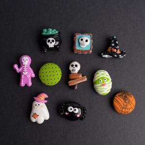 May include: A collection of Halloween-themed decorative items on a black surface. The items include a witch's hat, a cauldron with a skull, a mummy, a ghost, a spider, and a skull in a frame. The items are in various colors, including black, white, green, and orange.
