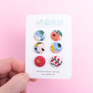 May include: Six pairs of nickel-free button earrings with floral patterns. The earrings are displayed on a white card with the text "aidille" and "Nickel Free Button Earrings www.aidille.com".