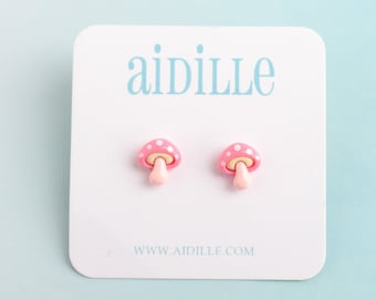May include: A pair of pink mushroom stud earrings with white polka dots, displayed on a white card with the brand name "aidille" in light blue. The earrings have a light pink stem and are set against a light blue background.