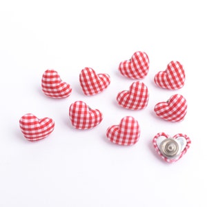 May include: A collection of red and white gingham heart-shaped fabric buttons scattered on a white surface. One button has a silver metal backing. These decorative buttons are ideal for craft projects.