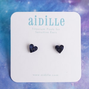May include: A pair of heart-shaped earrings with a galaxy-like design. The earrings are made of resin and have titanium posts for sensitive ears. The card reads "aidille Titanium Posts for Sensitive Ears" and "www.aidille.com".