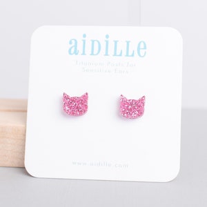 May include: Pink glitter cat stud earrings with titanium posts for sensitive ears. The earrings are displayed on a white card with the brand name "aidille" and the website address "www.aidille.com".