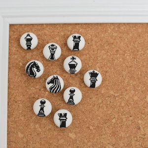 May include: A collection of white, round push pins with black chess piece designs, including a king, queen, rook, bishop, knight, and pawn. The pins are arranged on a corkboard, framed by a white border.