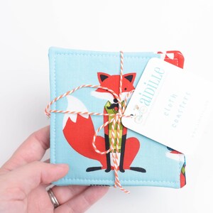 May include: A set of square cloth coasters tied with orange and white twine. Each coaster features a cartoon fox in red, white, and black on a light blue background. A white tag reads "aidille cloth coasters".