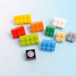 May include: A collection of colorful toy building blocks in various colors, including yellow, red, orange, green, blue, white, tan, and gray. A small black square with a silver button is also present. The blocks are scattered on a light blue surface.