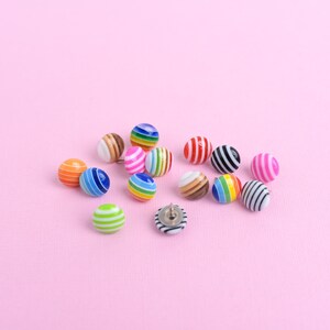 May include: A collection of small, round, dome-shaped buttons featuring various colorful striped patterns. Each button has a smooth, glossy finish with distinct horizontal or vertical lines in colors including blue, green, pink, orange, brown, and black.