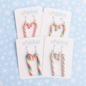 Peut inclure: Quatre paires de boucles d'oreilles en forme de cannes en sucre sur des cartes blanches. Les boucles d'oreilles sont de différentes couleurs, dont des rayures rouges et blanches, arc-en-ciel et rayures vertes et rouges. Le nom de la marque "aidille" est visible.