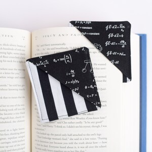 May include: Two black and white striped fabric corner bookmarks with white mathematical equations printed on them. The bookmarks are placed on an open book.