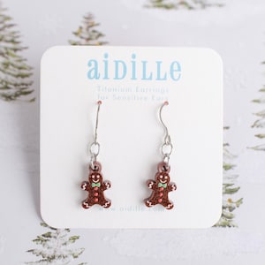 May include: A pair of gingerbread man earrings with silver hooks. The earrings are brown with red and green accents. The earrings are displayed on a white card with the brand name "aidille" in blue text.