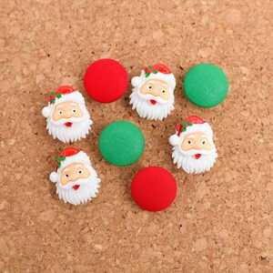 May include: A collection of Christmas-themed embellishments, including Santa Claus faces and fabric-covered buttons. The Santa faces are detailed with red hats, white beards, and rosy cheeks. The buttons are red and green.