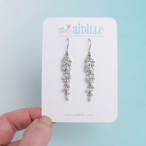 May include: Silver-toned dangle earrings with a cascading leaf design, adorned with clear crystals. The earrings are displayed on a white card with the brand name "aidille" and website address.