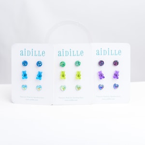 May include: Three sets of stud earrings on white cards. Each card features three pairs of earrings: a glittery stud, a gummy bear, and a heart. The earrings come in blue, green, and purple. The brand name "aidille" is printed at the top.