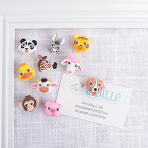 May include: A collection of colorful animal-shaped magnets, including a panda, donkey, tiger, duck, zebra, sheep, cow, monkey, pig, and dog. The magnets are affixed to a white framed board with a business card.