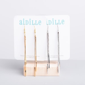 May include: Two pairs of long, dangling earrings on a white display card. The earrings are made of thin, linked chains. One pair is gold and the other is silver. The card has the text "aidille" printed on it in blue.