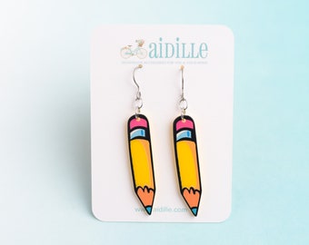 May include: A pair of dangle earrings shaped like yellow pencils with pink erasers and blue tips, suspended from silver-toned metal hooks. The earrings are displayed on a white card featuring the text aidille and a bicycle graphic.