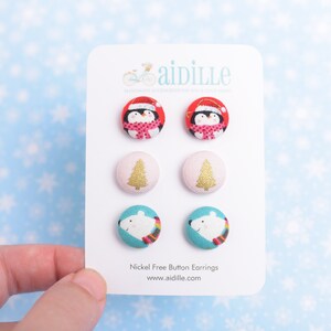 May include: A set of six nickel-free button earrings featuring three different Christmas designs: a penguin wearing a Santa hat, a Christmas tree, and a polar bear wearing a scarf. The earrings are displayed on a white card with the brand name "aidille" and the text "Nickel Free Button Earrings" and "www.aidille.com".